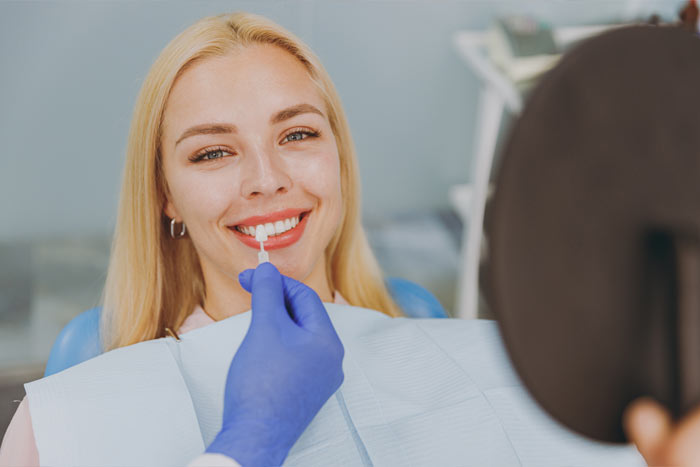 A smiling woman with blonde hair in a dental chair, receiving a cosmetic dental treatment from a dentist wearing blue gloves.