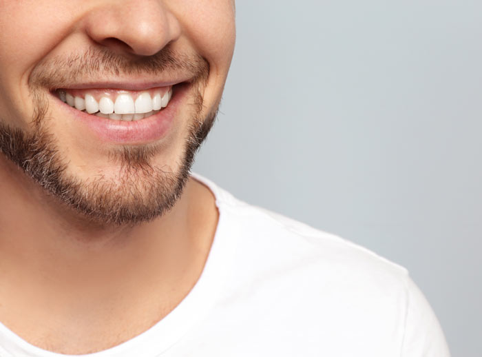 Close-up of a smiling man with a well-groomed beard, showcasing bright white teeth against a light gray background.