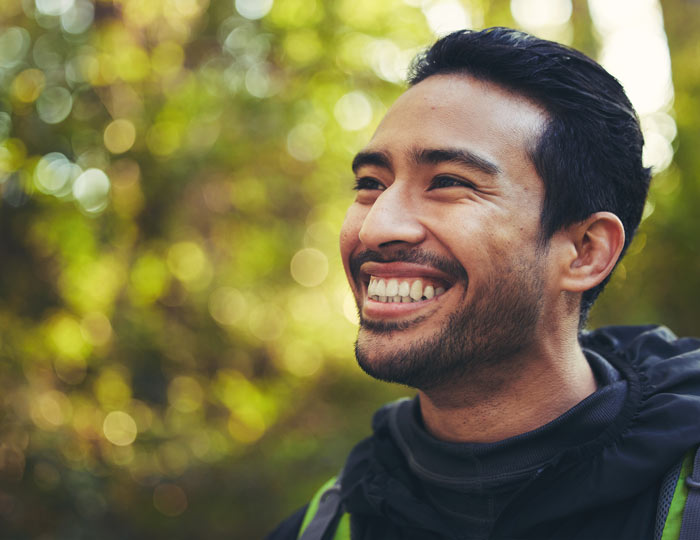 A smiling man with dark hair outdoors, surrounded by blurred greenery, exuding happiness and positivity.