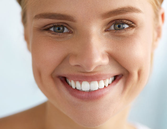 A joyful young woman smiling broadly, showcasing her bright white teeth and clear blue eyes, against a soft, light background, conveying a sense of happiness and confidence.