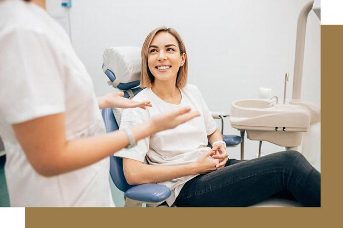 A smiling woman sitting in a dental chair, engaging in conversation with a dental professional, in a bright clinic setting.