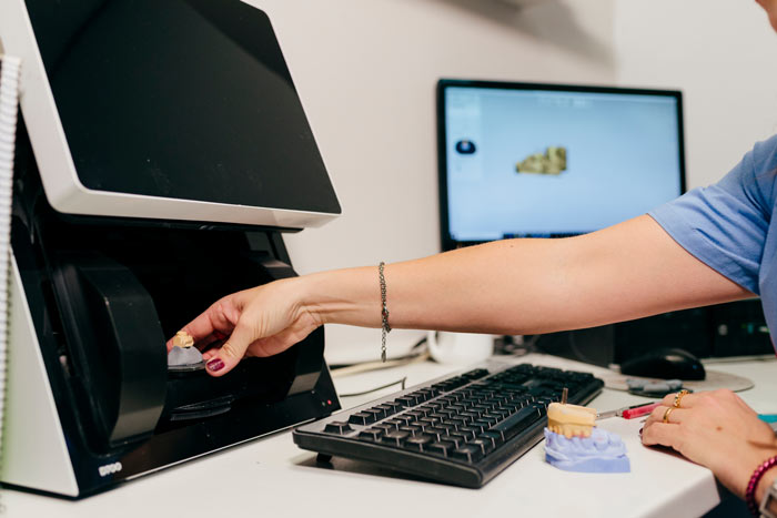 A person inserting a dental model into a 3D printer on a desk with a keyboard and computer monitor displaying a dental scan.