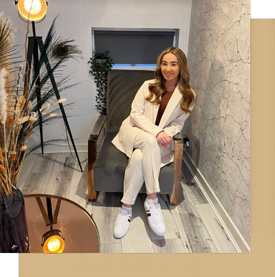 Young woman sitting in a stylish chair, wearing a light-colored blazer and sneakers, with decorative plants and modern furnishings in the background. Perfect for lifestyle and interior design themes.