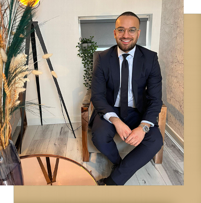 man sitting in a modern office environment, dressed in a navy suit with a white shirt and tie, smiling confidently, with a stylish table and decorative plants in the background, professional attire for business meetings.