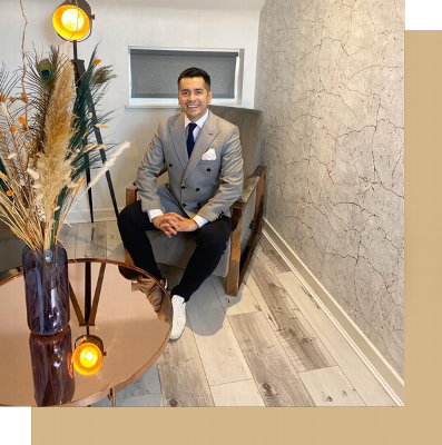 A well-dressed man sitting on a chair in a stylish interior, featuring a decorative vase with dried flowers and a modern side table with a lamp.