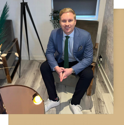 A man sitting on a chair, wearing a blue suit, white shirt, and green tie, with a stylish watch and white sneakers, posed confidently in an elegantly designed room with modern decor.