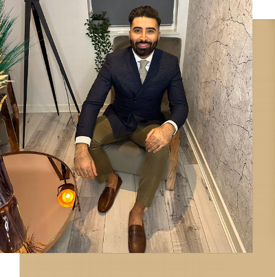 A well-dressed man sitting in a stylish lounge chair, wearing a navy blue blazer, light shirt, and beige trousers, with brown shoes. The modern interior features a decorative table, plants, and elegant decor.