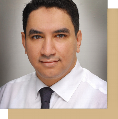 A professional man with short black hair wearing a white dress shirt and a dark tie, looking directly at the camera, against a softly blurred background.